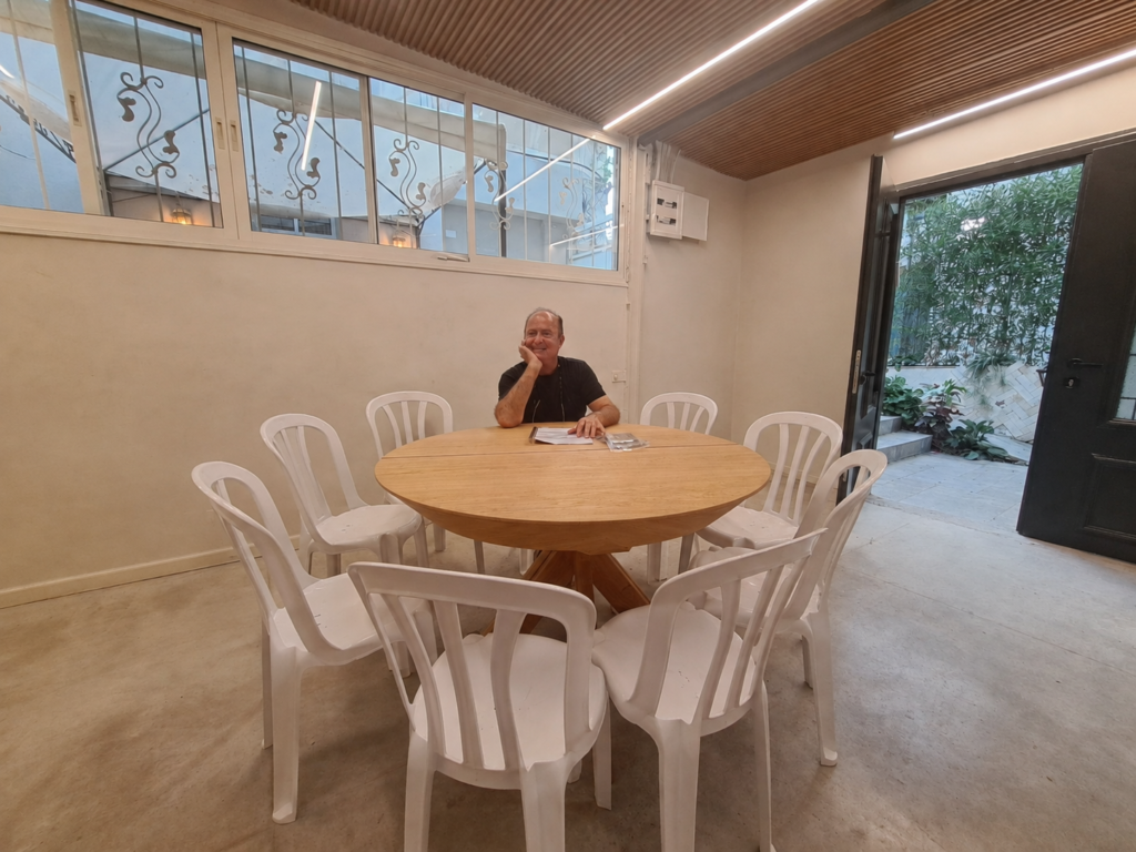 חנן חיים יושב ליד שולחן עגול מוקף כיסאות לבנים, רגע לפני מפגש בבית המאזן New Rise בתל אביב
